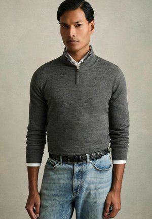 Hombre con el pelo corto y negro que lleva un jersey gris con cremallera sobre una camisa blanca y vaqueros azul claro sobre un fondo sencillo.