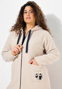 Sudadera de forro polar beige con cordones azul marino, cierre de cremallera, dos bolsillos delanteros y un parche en forma de corazón con un patrón de vaca en el bolsillo izquierdo.
