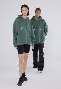 Πράσινα φούτερ με κουκούλα και μπροστινές τσέπες καγκουρό, φορεμένα με μαύρες σορτς ποδηλασίας. Παρουσιάζονται δύο μοντέλα, το ένα όρθιο μπροστά και το άλλο πίσω.