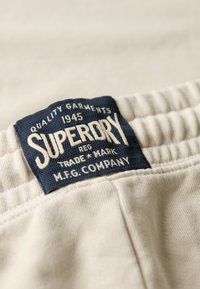 Beige stof met een gestructureerd oppervlak; heeft een blauw label met de tekst "SUPERDRY," waarop in witte letters kwaliteits- en merkinformatie staat.