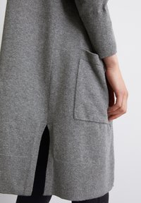 Grå stickad tröja med en sidofissur och en lapplomme. Mjuk textur, långa ärmar och ribbad nederdel.