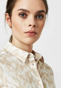 Beige Bluse mit abstraktem hellbraunem Muster, Knopfkragen und einem einzigen Knopfverschluss, aus einem glatten Stoff gefertigt. Sichtbares goldenes Creolen-Ohrgerät.