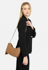 Borsa a tracolla marrone trapuntata con forma curvilinea, dotata di una catena dorata e logo in rilievo. Indossata sulla spalla sopra un blazer nero.