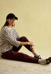 Gestreiftes beige-und-navy Oberteil, burgunderfarbene Cordhose, beige Socken mit Pünktchen und braune Schuhe. Schwarze Mütze getragen, sitzende Pose auf leichtem Teppich.