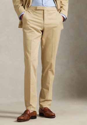 Homme portant un pantalon beige, une chemise bleu clair, un blazer beige, les mains dans les poches et des mocassins en cuir marron.