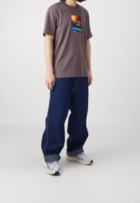 Brun grafisk t-shirt med en färgglad design på framsidan, tillsammans med löst sittande blå denimjeans och gråa sportskor.