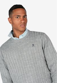 Maglione grigio a maglia con motivo a treccia, scollatura rotonda e piccolo logo ricamato sul petto, indossato sopra una camicia a righe azzurra chiara.