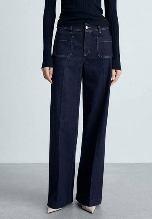 Femme portant un jean large bleu foncé avec des poches plaquées devant, associé à un haut noir côtelé à manches longues et des chaussures pointues beiges.