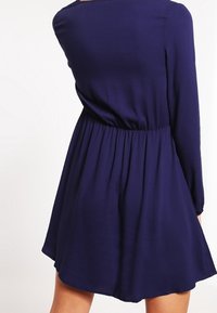 Robe bleu marine à manches longues, taille cintrée avec un élastique, et jupe évasée. Texture lisse avec un design simple et sans embellissements.