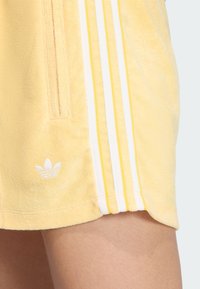 Žlté teplákové šortky z froté s bielymi a žltými pruhmi a malým bielym logom na leme. Obsahujú bočnú kapsu.