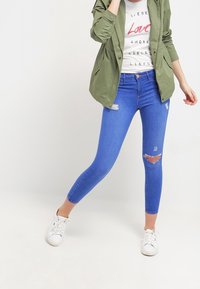 Blå smala jeans med slitna detaljer vid knät, matchade med en vit t-shirt med grafik och en grön jacka, samt vita sneakers.