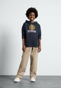 La felpa navy presenta una tasca frontale e un logo dorato; abbinata a pantaloni cargo beige e sneakers bianche. Texture morbide, design casual.