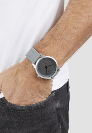 Gray silicone strap watch with a round black dial. Silver hardware with minimalist hour markers and hands. Worn on a wrist.