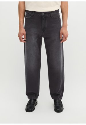 Mann trägt lockere schwarze Jeans aus Denim und schwarze Leder-Loafers, steht vor einem einfarbigen hellen Hintergrund.