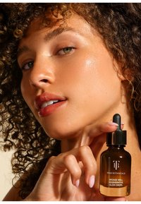 Bottiglia dropper in vetro marrone con tappo nero e dettagli dorati, etichettata "TRUE BOTANICALS BRONZE WELL REGENERATIVE GLOW DROPS." Texture della pelle umida.
