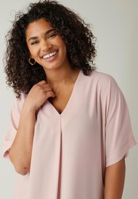 Jasnoróżowa bluzka z krótkim rękawem i dekoltem w kształcie litery V, ozdobiona plisowanym przodem. Wykonana z miękkiego, gładkiego materiału o luźnym kroju.