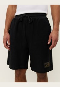 Shorts en tricot noirs avec une finition texturée, taille élastique et cordon de serrage. Présente un petit patch logo doré sur la jambe inférieure.