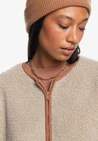 Beige fleecejacka med en brun dragkedja och krage, bärs över en matchande brun topp. Tillbehöret inkluderar ett enkelt kedjearmband.