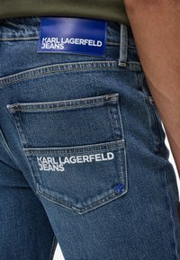 Sötétkék farmernadrág, texturált felülettel, hátulján kék bőrcímkével, rajta fehér "KARL LAGERFELD JEANS" felirattal.