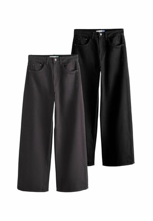 Person trägt schwarze Jeans, graues Hemd und beige Jacke mit einer Hand in der Tasche, zugeschnitten von der Taille bis zur Mitte des Oberschenkels.