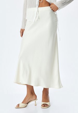 Jupe longue en satin blanc fluide avec une texture lisse, ourlet évasé et taille ajustée, associée à des sandales à talons ouverts de couleur claire.