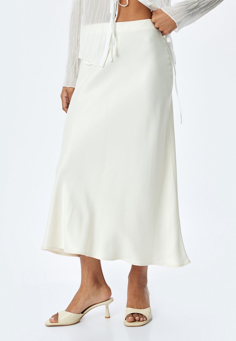 Jupe longue en satin blanc fluide avec une texture lisse, ourlet évasé et taille ajustée, associée à des sandales à talons ouverts de couleur claire.