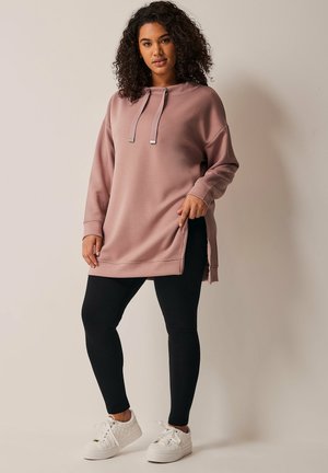 Rosa oversized hoodie med snoretræk i hætten, sidesplidser, parret med sorte leggings og hvide sneakers på en neutral baggrund.