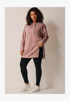 Sudadera rosa de gran tamaño con capucha ajustable, cortes laterales, combinada con leggings negros y zapatillas blancas sobre un fondo neutro.