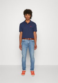 Polo navy con logo rosso, abbinato a jeans blu chiaro e sneakers arancioni. L'outfit è completato da una cintura in pelle marrone per ulteriori dettagli.