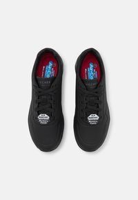 Baskets de sport noires avec un design à lacets, en matériau synthétique texturé, avec des perforations et des semelles intérieures rembourrées étiquetées "Antidérapant" et "Mémoire de forme."