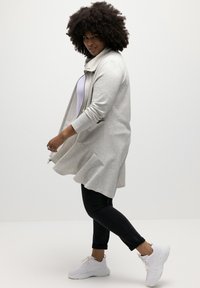 Chaqueta larga gris claro con cuello, cierre de cremallera y dobladillo acampanado, combinada con jeans negros y zapatillas blancas. Textura de tela suave visible.
