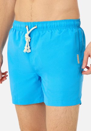 Mand iført lyseblå badeshorts med hvid snorknude og lille sideetiket, torso og øvre ben synlige.