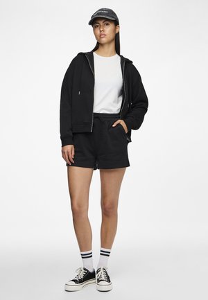 Jonge vrouw die een zwart vest met rits draagt, zwarte korte broek, wit T-shirt, zwarte pet, witte sokken met zwarte strepen en zwarte sneakers, staand.