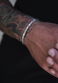 Metallist käevõru, millel on graveeritud sõnum, hõbedane, ümmargune kuju, kantud pruuni nahaga randmel, koos nähtavate tätoveeringutega.
