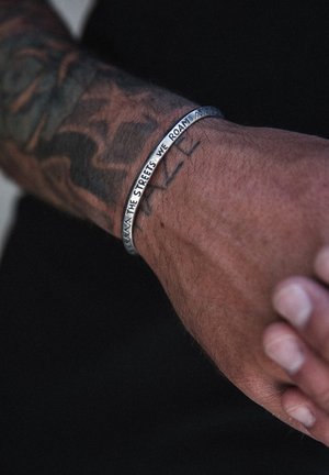 Metal bracelet with an engraved message, silver-colored, round shape, worn on a tanned wrist, alongside visible tattoos.