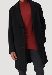 Manteau en laine noir avec un col à revers cranté, six boutons et des poches latérales, porté sur un pull rouge côtelé, sur un fond neutre.