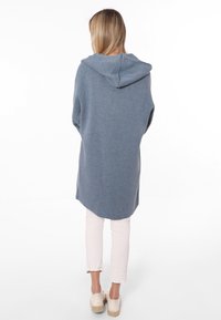 Długi niebieski hoodie z teksturowanej tkaniny, z kapturem, opadającymi ramionami i lekko zaokrąglonym dołem. W zestawie z jasnymi spodniami.