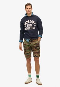 Marineblauwe sweatshirt met "Superdry"-logo, camouflage cargoshorts, witte sneakers en groen gestreepte sokken, gecombineerd met een lichtblauw overhemd.