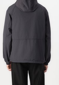 Homme portant une veste à capuche sombre unie avec des poignets élastiques, vu de dos sur un fond blanc.