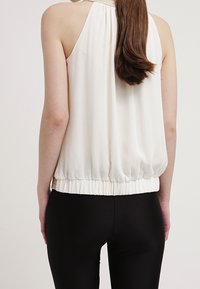 Haut sans manches crème avec une taille froncée et une texture lisse. Vue de dos montrant une silhouette fluide avec un léger éclat.