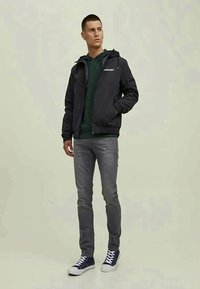 Jovem está de pé, usando uma jaqueta preta com capuz sobre um hoodie verde escuro, jeans cinza e tênis de cano alto azul-marinho, contra um fundo simples.