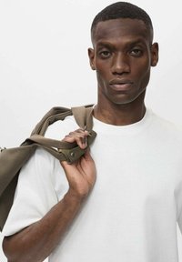 Tricou alb cu mâneci scurte și croială lejeră. O persoană ține curelele de un sac de culoare verde măslin pe umăr. Fundalul este simplu.