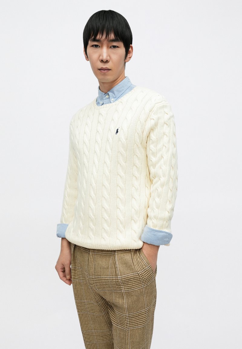 Cream cable-knit sweater layered over a light blue collared shirt, paired with beige plaid trousers. The look features textured patterns and fitted style.