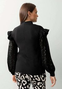 Schwarzes Oberteil mit kurzen, schlichten Ärmeln und langen, mit Pailletten besetzten Ärmeln; kombiniert mit schwarz-weißen gemusterten Hosen. Sichtbarer Reißverschluss auf der Rückseite.