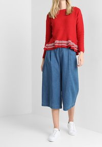 Red knitted sweater with ribbed hem and white stripes, paired with loose blue culottes and white sneakers. Smooth textures throughout.