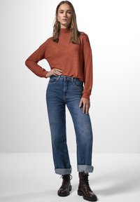 Top a maniche lunghe color ruggine con collo alto, abbinato a jeans azzurri con risvolti e stivaletti alla caviglia marroni con lacci.