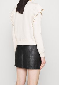 Sudadera beige con detalles de hombros fruncidos, puños y dobladillo acanalados, combinada con una mini falda de cuero negra, mostrando texturas suaves y líneas limpias.
