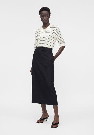Γυναίκα που φοράει λευκή πλεκτή μπλούζα με κοντά μανίκια και ριγέ κοψίματα, μαύρη midi φούστα με ψηλή μέση και μαύρα σανδάλια με τακούνι, πόζα με το ένα χέρι στην τσέπη.