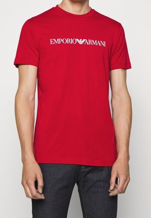 Homme portant un t-shirt rouge Emporio Armani avec un logo blanc, associé à un jean bleu foncé, debout devant un fond uni.