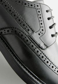 Chaussure en cuir noir avec un design perforé, des coutures décoratives et une texture lisse, dotée de cinq œillets pour les lacets.